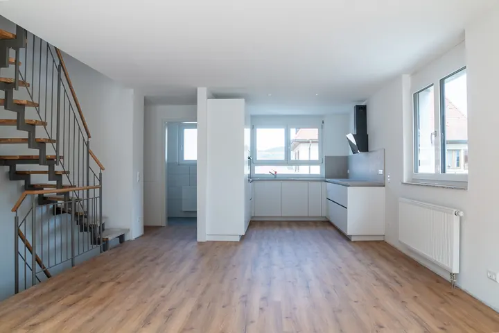 Offener Wohnraum mit Stahltreppe links, weißer Einbauküche in der Mitte und Seitenfenster rechts